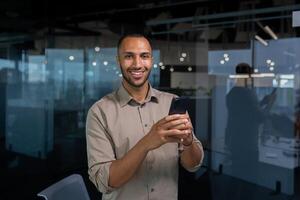 Portrait young hispanic man inside office man in a casual shirt is smiling and looking at the camera, a businessman is holding a smartphone in his hands, typing a message on the phone, making a call. photo