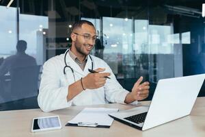 Online reception at the hospital. Young handsome hispanic doctor talking to a patient on a call from a laptop. Sits in the office at the desk, talks to the camera, explains and waves his hands photo