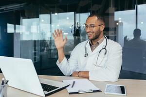 Happy hispanic doctor smiling and talking on call man in white coat and stethoscope using laptop for remote online consultation smiling and waving hello. photo