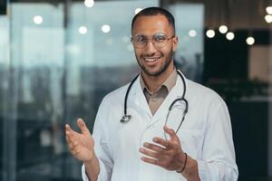 Close-up photo of hispanic doctor smiling and looking at web camera talking to colleagues and patient on call, man in medical coat and stethoscope working inside clinic office.
