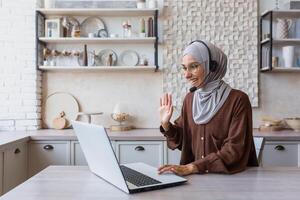 Woman in hijab working remotely from home sitting in kitchen using laptop and headset for call, businesswoman smiling and talking to customers remotely customer support service online tech photo