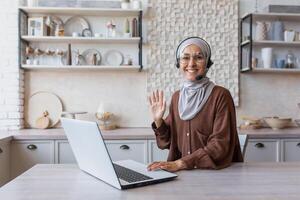 Young Muslim woman in hijab and headset, blogger doing remote online broadcast, communicating via call. Sitting at home in the kitchen at the laptop. A smiling woman waves hand at the camera. photo
