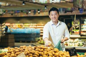 Portrait of an Asian shopper in the grocery department of a supermarket, a man chooses vegetables and buys potatoes, smiling and looking at the camera photo