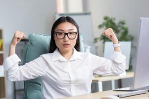 Successful Asian business woman looking at the camera and holding her hands up, a gesture of super power and success, an employee in the office works at a computer. photo