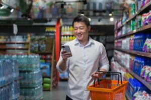 Experienced mature Asian shopper in supermarket, man smiling and reading message on smartphone, makes purchases and chooses products from various shelves photo