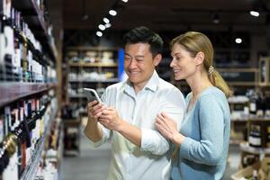 Young diverse couple man and woman in liquor department choosing wine, smiling and happy using smartphone to identify wine, using app to scan products photo