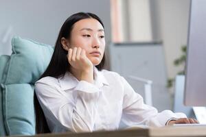 Boring and monotonous work. A young beautiful Asian business woman, an office worker sits at a desk at a computer, is sad, bored, performs monotonous work. He holds his head with one hand. photo