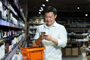Happy Asian shopper in supermarket using smartphone app to select and scan wine in store, man among wine shelves in liquor department photo