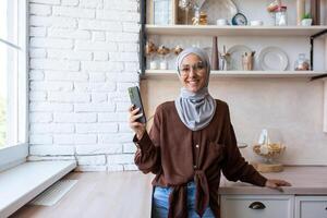 A young Muslim woman in a hijab is standing at home in the kitchen by the window and using the phone. Smiling, she looks at the camera. photo