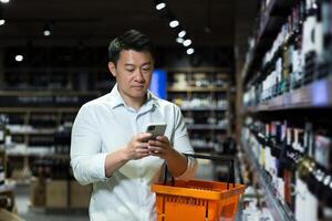 A young man, an Asian businessman chooses wine in the alcohol section of a supermarket, holds a phone in his hands, searches for the name of wine, composition, production in the search engine. photo