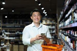 Asian man in supermarket, uses phone and app to buy and choose wine in store, looks at camera and smiles photo