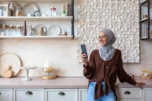 Smiling Muslim young woman in hijab standing in kitchen at home and using mobile phone, reading news, checking social media, watching videos, photos, searching for information. photo