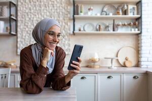 Close up photo. Smiling Muslim young woman in hijab standing in kitchen at home and using mobile phone, reading news, checking social media, watching videos, photos, searching for information. photo