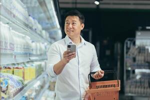asian man shopper walks around the supermarket with a shopping cart looking at smart phone browsing smartphone in hands search ,goods or products on the list customer purchaser male in a grocery store. photo