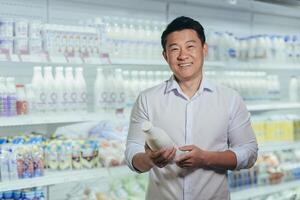 Portrait of happy and satisfied asian shopper in supermarket in dairy department, man holding bottle of milk smiling and looking at camera near refrigerators photo
