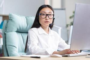 Portrait of a young Asian woman in headphones with a microphone. Works as a consultant in a call center. Sitting at the table in glasses at the computer, looking at the camera, smiling. photo
