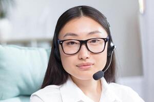 Close-up portrait photo of Asian female call center worker and tech support worker, looking at camera and smiling, using headset for call.