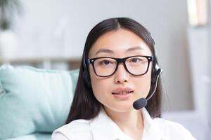 Portrait of a young Asian woman in headphones with a microphone. Works as a consultant in a call center, answers calls. Sitting at the table in glasses at the computer,looking at the camera, smiling. photo