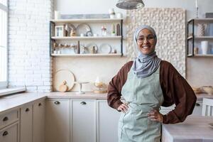 A young Muslim woman food blogger, trainer, coach, chef in hijab standing at home in kitchen at table in apron, hands on waist, posing, smiling and looking at camera. Conducts online courses, lessons. photo
