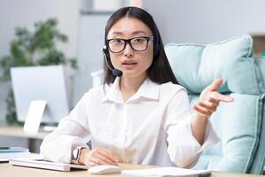 Beautiful Asian female bank support technician looking into camera and talking on call, using headset, explaining information to customers, helpline. photo
