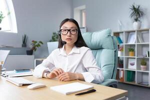 Asian woman in headphones with a microphone, sitting in the office at the desk conducting an online webinar, explaining, consulting, telling, gesturing with her hands. He looks at the camera, smiles. photo