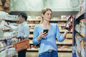 A beautiful young blonde woman is standing in a supermarket. She is holding a phone and a packet of breakfast cereal. Chooses, buys, reads the label. Shoppers in the background. photo