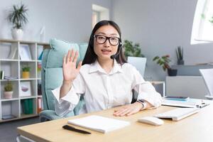 Young Asian call center worker looking at camera smiling waving greeting gesture using headset for call. photo