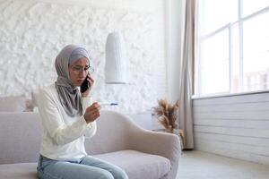 Upset and disappointed young arab woman in hijab sitting on sofa at home and talking on the phone. Holds a quick and one-time pregnancy test with a negative result. photo