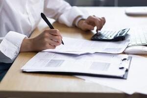 Close-up photo. Beautiful hands of a young woman in a white shirt. They write documents with a pen, calculate on a calculator at the table in the office, where there is a computer, a cup of coffee. photo