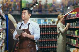 Two workers in a supermarket in the grocery department arrange products, an Asian man with a tablet computer and a woman in aprons among the products on the shelves and racks photo