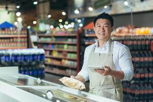Portrait of Asian store manager, man with tablet checking expiration date of products, seller smiling and looking at camera near refrigerator in supermarket photo