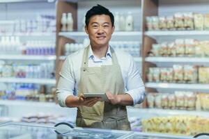 portrait of happy asian male small business owner standing in grocery store behind counter. Male manager with a tablet looking at the camera. friendly administrator food market. worker in apron. photo
