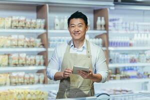Portrait of a young handsome Asian man, shop owner, a supermarket worker. Standing in an apron in the dairy department of the store with a notebook in his hands, looking at the camera, smiling. photo