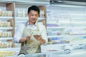 Internship at work. A young man, an Asian stager, an assistant works in a supermarket. Standing in an apron in the dairy department near the refrigerators, writing down the product in a notebook. photo