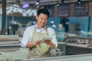 Asian grocery store manager salesman in apron using digital tablet counting goods in supermarket worker employee doing inspection of products regular inventory to control goods online order. photo