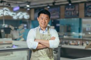 portrait of handsome asian man salesman in apron standing and looking at camera in grocery store supermarket. Male worker seller or small business owner. indoor. food, groceries market Cheerful smile photo