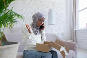 Problems with home delivery. Young muslim woman in hijab sitting on sofa and talking on the phone, holding a box on her lap and spreading her hands worriedly. I received the wrong parcel, damaged. photo
