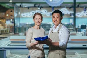 Asian male seller in supermarket recommending product to female buyer, couple looking at camera and smiling satisfied with shopping service near refrigerators in store. photo