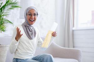 Young female Muslim student in hijab is sitting on the sofa at home and reading the letter received from the university with the results of the exam, interview, job acceptance. Happy looking at the camera. photo
