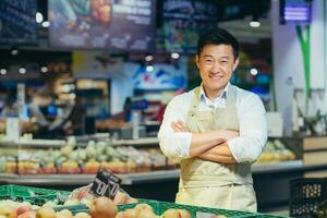 Portrait of a handsome young Asian man, owner and director, businessman of a store, supermarket. Standing in an apron in the vegetable and fruit department, arms crossed, looking at the camera, smiling. photo