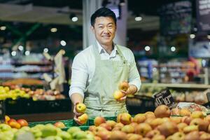 Waist up portrait of a handsome Asian handsome man working in a supermarket and smiling at the camera. Standing in a work apron near separated vegetables and fruits. He holds apples in his hands. photo