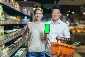 Portrait of a happy Asian couple of consumers, shoppers in a supermarket or grocery store, looking at the camera, smiling. are holding a phone and showing a green chromakey screen photo