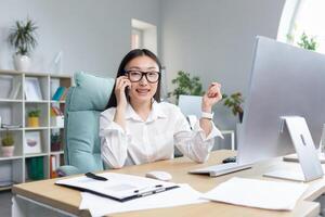 Beautiful asian office worker secretary working in modern office talking on the phone smiling and looking at camera. photo
