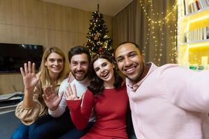 Diverse friends celebrating christmas and new year together, group of people having dinner at home sitting on sofa near Christmas tree, taking selfie photo and talking with friends on call using phone.