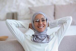 Muslim young business woman in hijab and glasses sitting on sofa at home with hands behind head and resting, smiling and satisfied with successful work done and finished. Smiling at the camera. photo