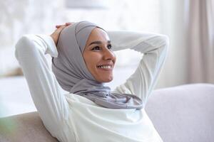 Close-up photo. Smiling arab young business woman in hijab sitting on sofa at home with hands behind head and resting, smiling and satisfied with successful work done and finished. photo