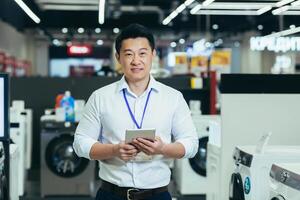 Happy Asian manager shop manager salesman smiling and looking at camera, man selling large household electronic appliances, using tablet computer in supermarket photo