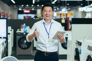 Successful sales consultant in electronics supermarket, Asian man working at home appliances store smiling and looking at camera, man holding tablet portrait and showing thumb up recommending photo