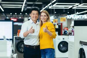 Satisfied diverse couple of shoppers Asian man and woman smiling and looking at camera, satisfied and positive and recommending store hold thumbs up, shoppers in electronics store photo
