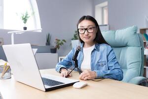 Podcast recording, interview. Young beautiful Asian woman sitting at a table with a microphone and a laptop. Talks, tells. Makes an audio recording of a podcast, gives an interview via call. photo